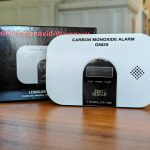 Vorderansicht des Kohlenmonoxid-Warnmelders GS828 mit Display, Test-Silence-Taste und Verpackung im Hintergrund auf Holztisch