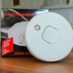 Weißer Rauchwarnmelder mit 10-Jahres-Lithium-Batterie und VdS-Prüfung vor seiner Verpackung mit der Aufschrift Smoke Alarm