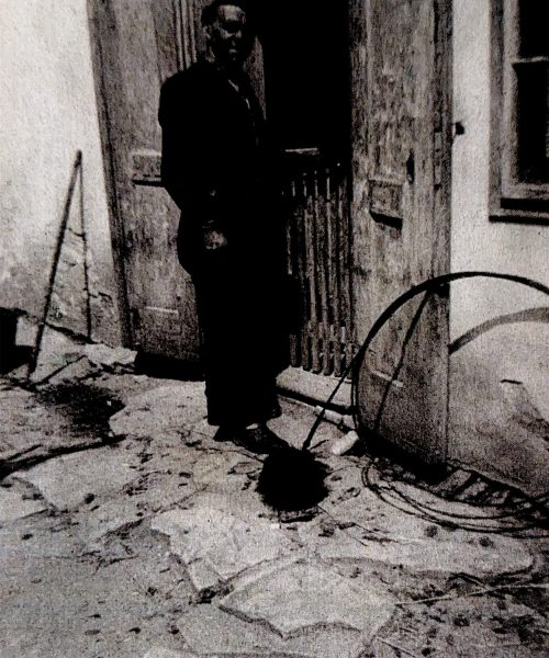 Schwarzweißfoto eines Rauchfangkehrers mit Werkzeug vor einem Haus, aufgenommen während seiner Arbeit am Kamin