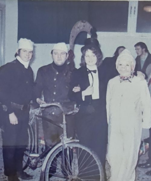 Altes Farbfoto mit Rauchfangkehrern in traditioneller Uniform, begleitet von festlich gekleideten Personen und einem alten Fahrrad, aufgenommen bei einer Feier oder Veranstaltung